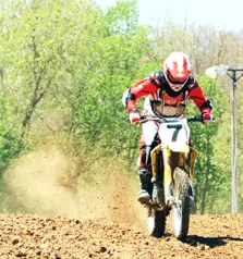 Dirtbike racer wearing red gear speeding on a dirt track with trees in the background - helmet label 