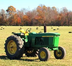 Green tractor in a hay field with fall foliage in the background 