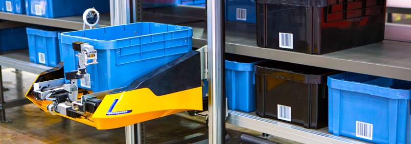 Automated yellow warehouse robot lifting a blue plastic bin with a printed barcode label applied to the surface.